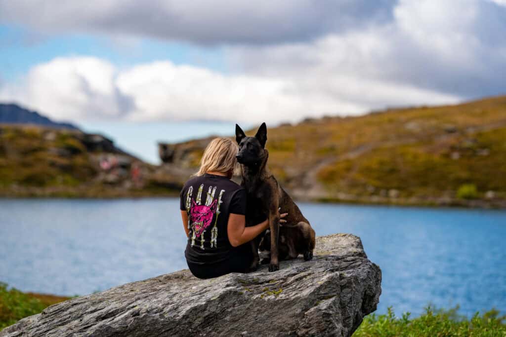 Forget Me Not Dog Training - A person with long blonde hair in a black T-shirt sits on a rock by a lake in South Carolina, next to a large brown and black dog, under a cloudy sky.