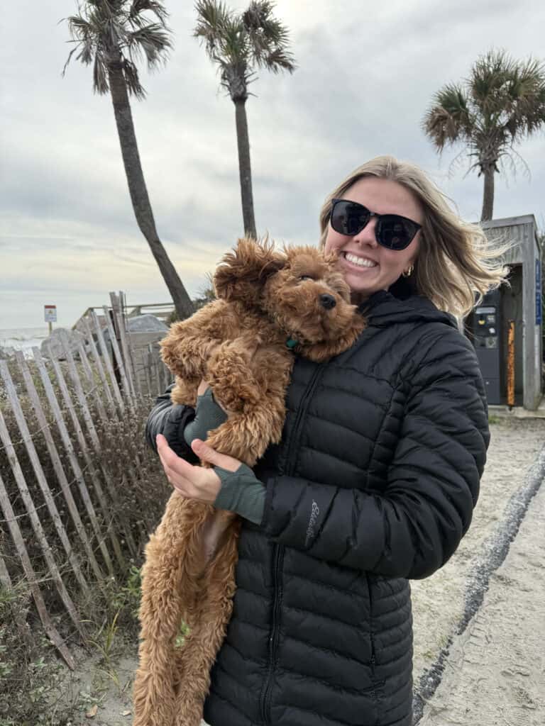 Forget Me Not Dog Training - A person in a black jacket and sunglasses holds a fluffy puppy on a South Carolina beach with palm trees swaying in the background.