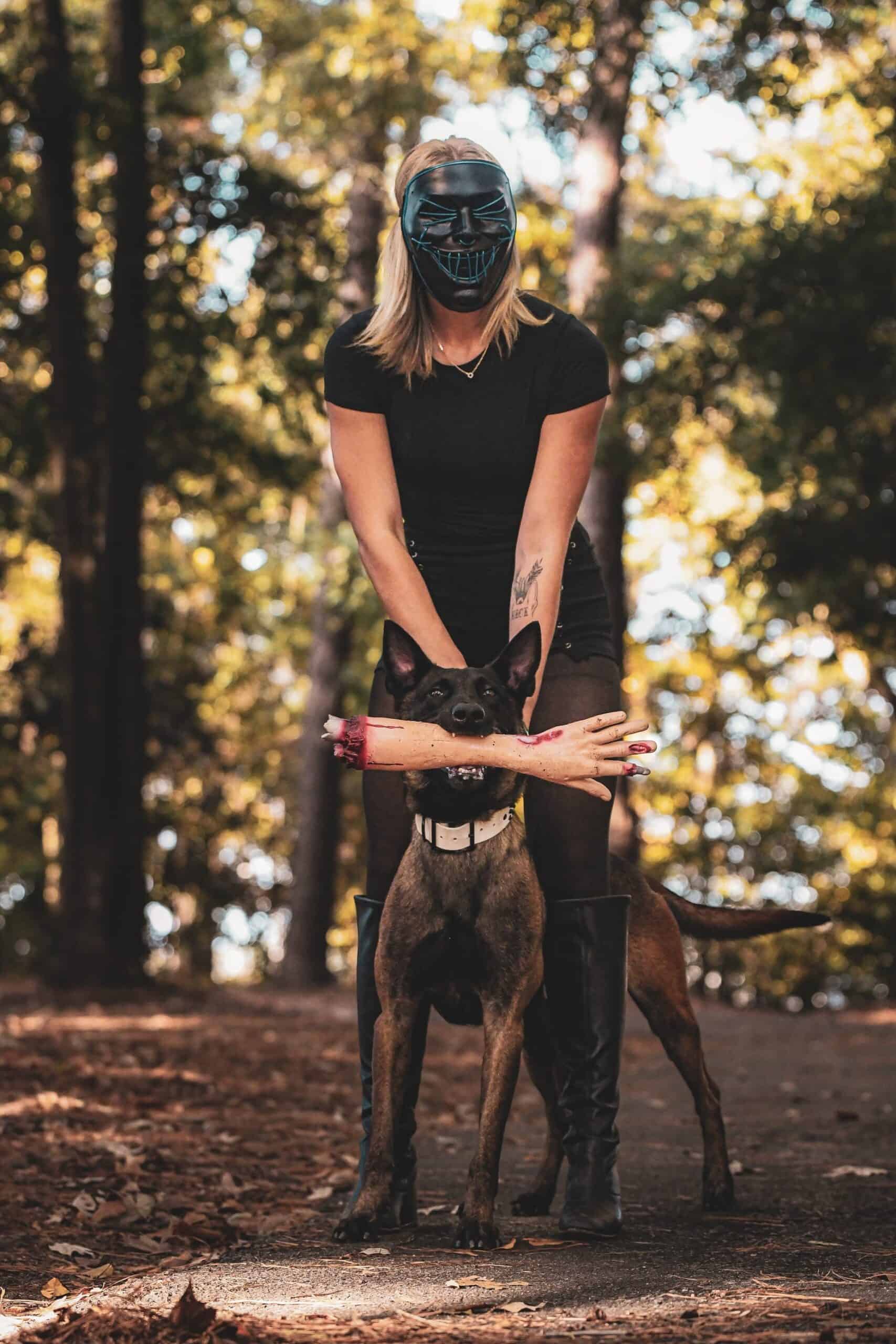 Forget Me Not Dog Training - A woman wearing a mask stands behind a dog holding a fake severed arm toy in its mouth on a wooded path.